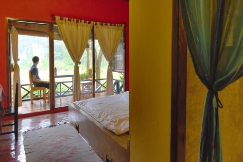 Un dormitorio con una cama y una persona sentada en una silla en un balcón en Mandala Ou Resort, en Nongkhiaw