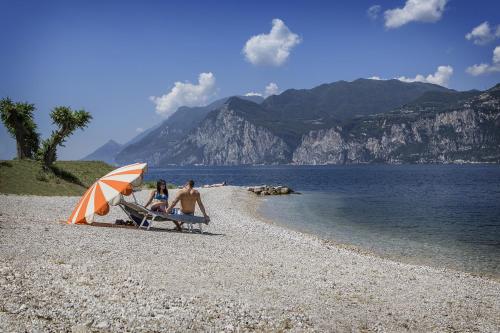 two people sitting on a beach with an umbrella at Ambienthotel PrimaLuna 3S in Malcesine