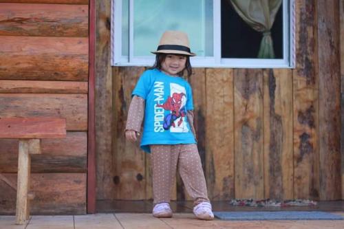 a little girl wearing a hat standing in front of a building at ไร่นิธิสุนทร nithisoonthon farmstay in Mae Hong Son