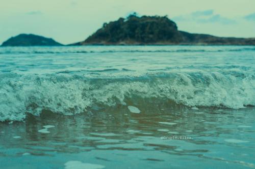 a small wave in the ocean with an island in the background at Pousada Acqua Bella PISCINA CLIMATIZADA in Bertioga