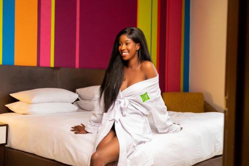a woman in a white dress sitting on a bed at Ibis Styles Accra Airport in Accra