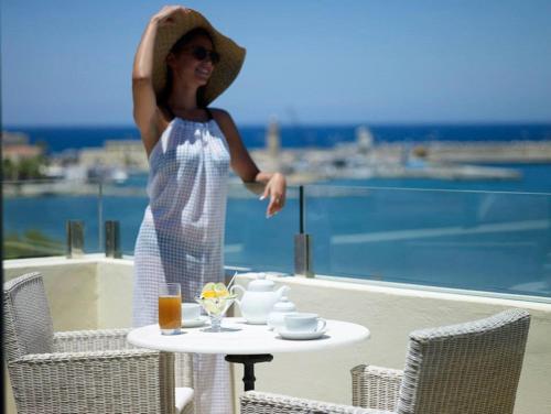 una mujer con un sombrero de pie en un balcón con una mesa en Kyma Suites Beach Hotel, en Rethymno