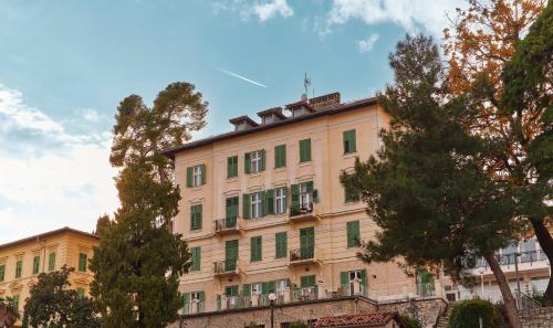 un edificio abbronzato con finestre e alberi con persiane verdi di Hotel Lovran a Lovran (Laurana)