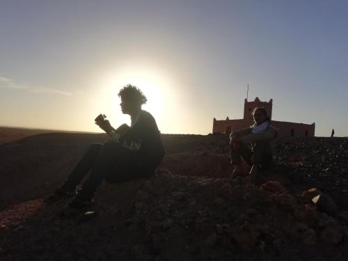 two people sitting on a hill with the sun in the background at Bivouac Les Nomades & Foum zguid to chegaga tours in Foum Zguid