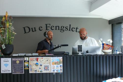 deux hommes sont assis sur un bureau avec une table et un ordinateur portable dans l'établissement Fengselshotellet, à Arendal