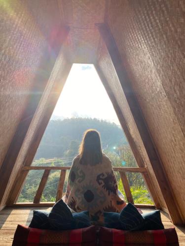 a woman sitting on a bed looking out of a window at Monsonkeang Camping view ม่อนสันเกี๋ยง in Mae Chaem