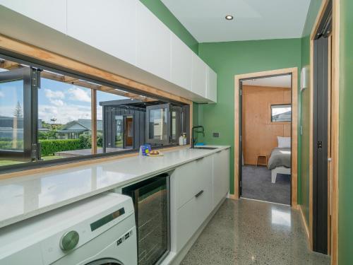 a kitchen with green walls and white cabinets and windows at The Little Bach at Whangapoua in Whangapoua