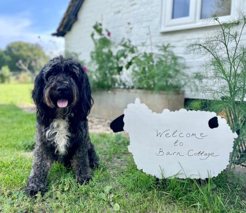 un perro negro sentado en la hierba con una señal de bienvenida en Barn Cottage -Westerlands en Graffham