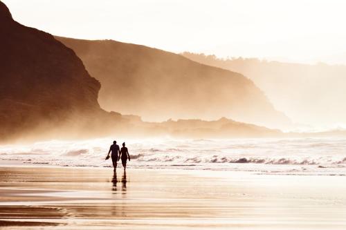 two people walking on a beach with a surfboard at Oceanfront Home Retreat w Breathtaking Views of Fitzgerald Marine Reserve in Moss Beach