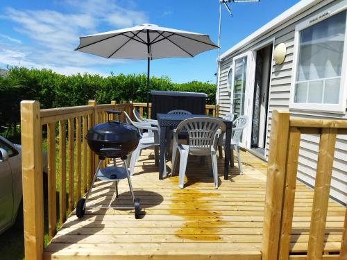 una terraza de madera con parrilla, sillas y sombrilla en Mobil home-Les Lins Bleus, en Biville-sur-Mer