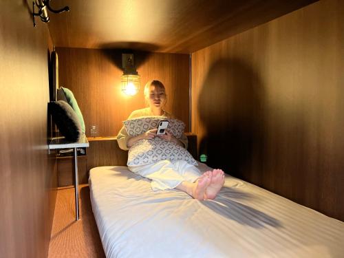 横浜市にあるHARE-TABI SAUNA&INN Yokohamaの携帯電話を見ながらベッドに座っている女性