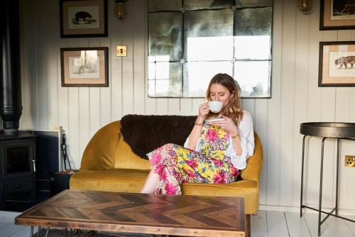 Eine Frau, die auf einer Couch sitzt und eine Tasse Kaffee trinkt. in der Unterkunft Hog Deer Creek - At Port Lympne Reserve in Hythe