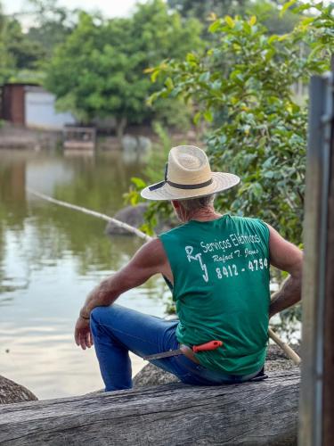 a man in a hat sitting on a dock with a boat at Seu Refúgio Rural Piscina Privativa e Natureza in Tubarão
