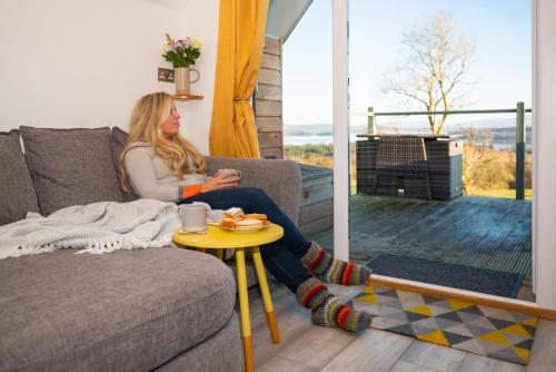 a woman sitting on a couch with a cup of coffee at Bonnie Barns - Adults Only Luxury Lodges with hot tubs in Luss
