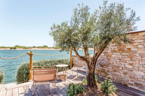 a patio with chairs and a tree next to the water at Relais et Châteaux Thalasso & Spa Ile de la Lagune in Saint-Cyprien