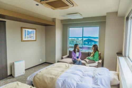 two women sitting in a room with a bed and a window at Ise-Shima Ohtaya Healing Stay in Toba