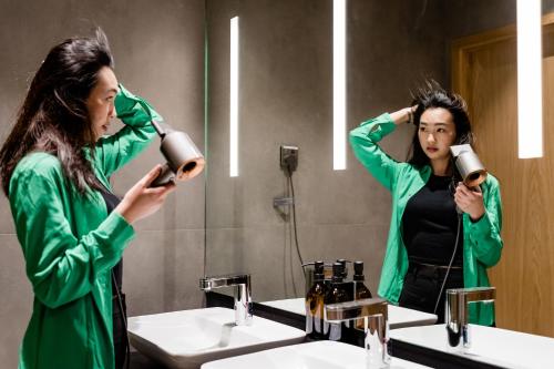 a woman drying her hair in front of a bathroom mirror at Sleepover Doha North Node - formerly sleep 'n fly in Doha