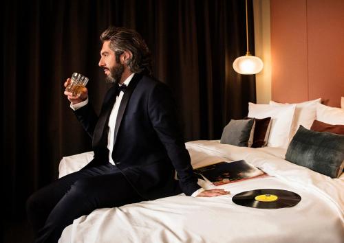 a man in a suit sitting on a bed holding a glass at Design-Hotel JAMS in Munich