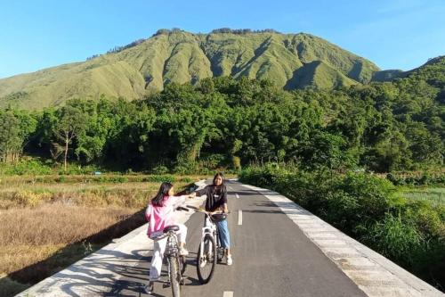 zwei Frauen reiten Fahrrad auf einer Straße mit Bergen im Hintergrund in der Unterkunft Sada 360 homestay in Sembalun Lawang