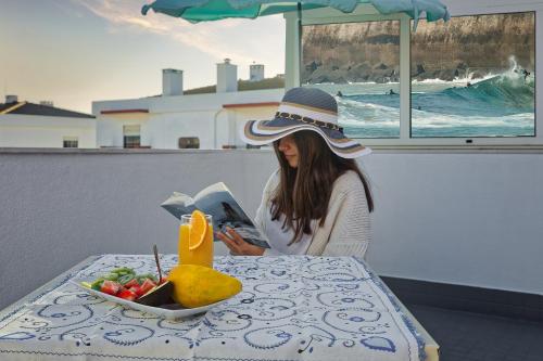kobieta w kapeluszu siedząca przy stole czytająca książkę w obiekcie Alojamento Santo António w mieście Peniche