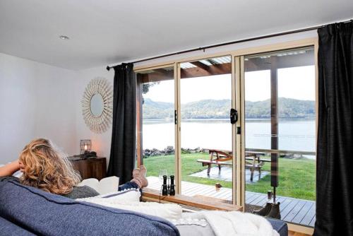 a woman sitting on a couch looking out at a large window at Shack on the Bay - A Lovely Bayside Cabin in Strahan