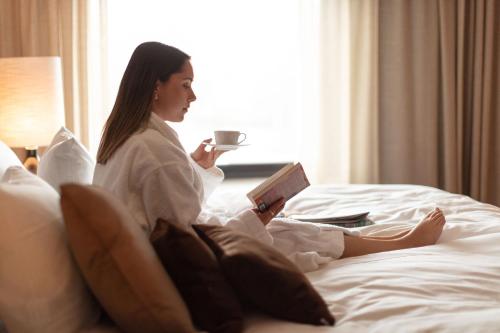 a woman sitting on a bed reading a book and holding a cup at Billingehus in Skövde