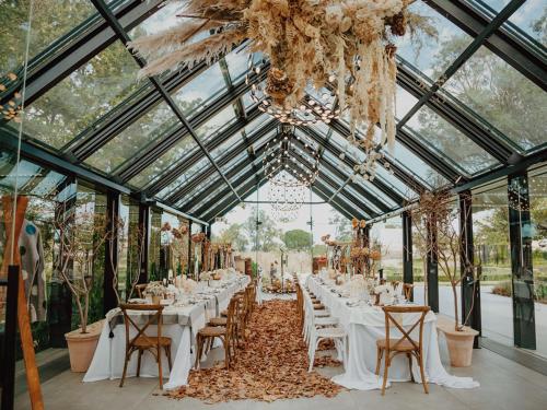 a long table with white tables and chairs in a greenhouse at The Hazendal Hotel in the Stellenbosch Winelands by NEWMARK in Stellenbosch