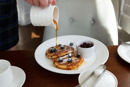 a person pouring syrup on a plate with blueberries and pancakes at The Roseate Villa Bath in Bath