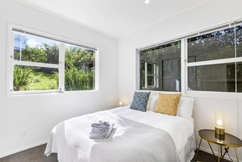 a white bedroom with a bed and two windows at Coastal Calm is near to the Beach with BBQ in Auckland