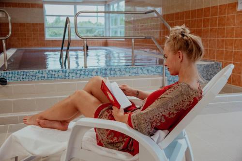 uma mulher sentada numa cadeira a ler um livro em frente a uma piscina em Arkánum Hotel em Balatonmáriafürdő