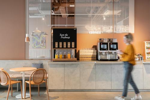 a woman walking past a coffee shop with a counter at ibis budget Gent Centrum Dampoort in Ghent