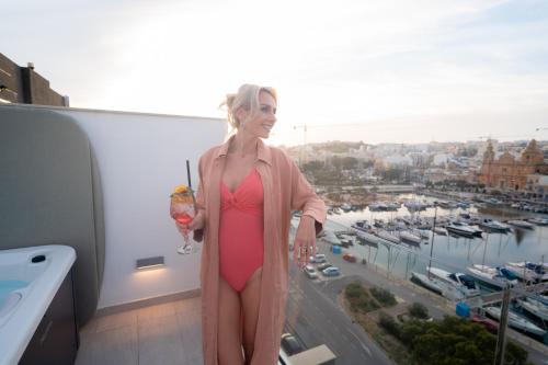 a woman in a pink dress standing on a balcony with a drink at The Address Seafront Suites with Hot Tub in Pieta