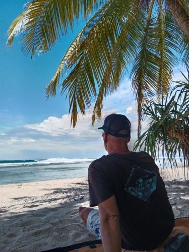 a man sitting on the beach under a palm tree at Rumah Nalu surf camp in Krui