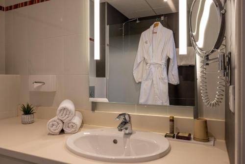 a bathroom sink with a white robe hanging in a mirror at Hôtel Val-Vignes Colmar Haut-Koenigsbourg, The Originals Relais in Saint-Hippolyte