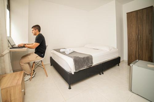 a man sitting on a chair next to a bed with a laptop at Descansa y Explora Habitación Privada en Medellín C in Medellín