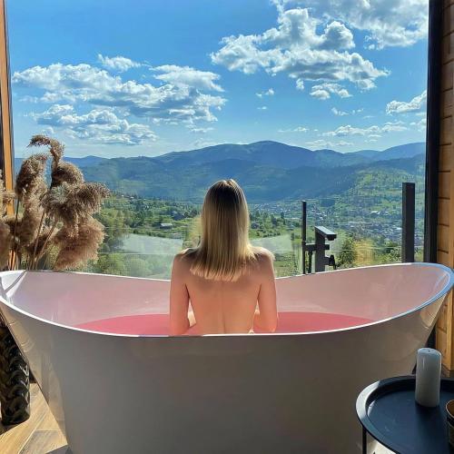 a woman sitting in a bath tub with a view at Under Sky Slavske - Котеджі з Теплим басейном ,чаном-джакузі, панорамною ванною та PlayStation 5 in Slavske