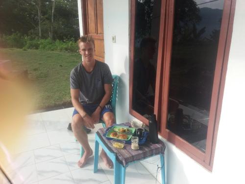 un hombre sentado en un porche con una mesa con comida. en Rice field guest house en Kelimutu