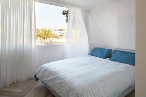 Cette chambre blanche dispose d'une fenêtre et d'un lit avec des oreillers bleus. dans l'établissement Luxury SeaSide DeShalit Square, à Herzliya