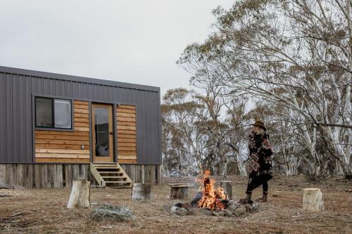 a man standing by a fire in front of a cabin at Big Yard Escapes - Two Off-Grid Tiny Houses on the Mowamba River in Jindabyne
