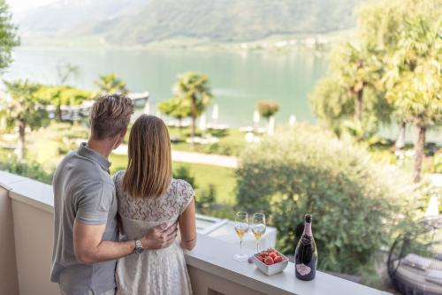 Ein Mann und eine Frau stehen auf einem Balkon mit Weingläsern. in der Unterkunft Parc Hotel Am See in Kaltern