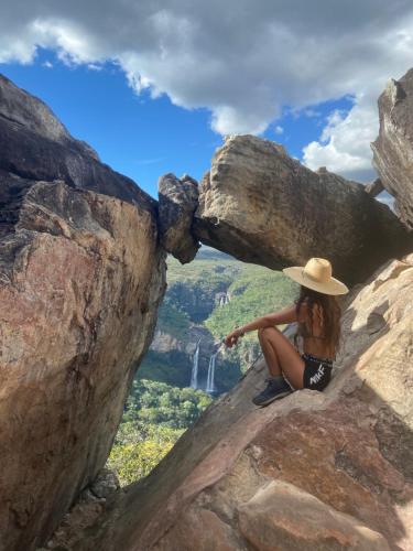 a woman in a hat sitting on a rock with a waterfall at Pousada/Camping Mirante Serrano in Alto Paraíso de Goiás