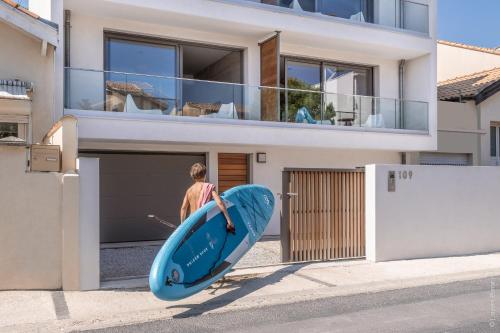 un hombre sosteniendo una tabla de surf azul delante de una casa en Superbe T2 neuf haut de gamme, climatisé, parking gratuit, 2eme ligne balcon, jardin en Carnon-Plage
