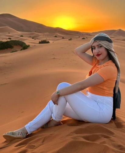 a woman sitting in the desert with the sunset in the background at Merzouga Top Luxury Camp in Merzouga
