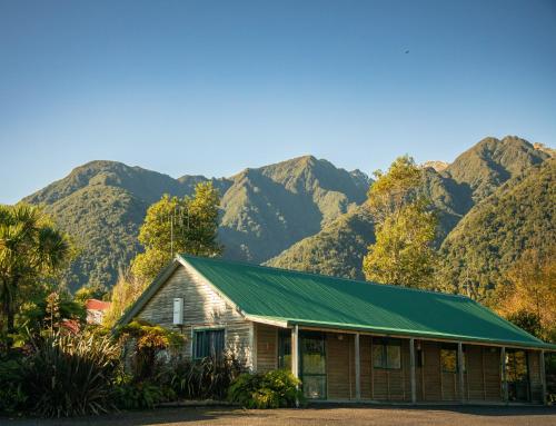 a house with a green roof with mountains in the background at Rainforest Motel in Fox Glacier