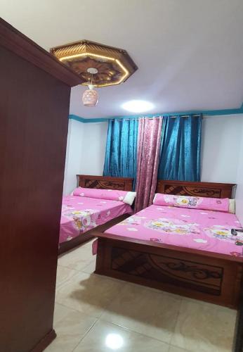two beds in a room with pink sheets and blue curtains at M&H Apartment in Dumyāţ al Jadīdah