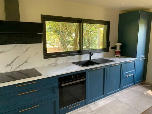 une cuisine avec des armoires bleues, un évier et une fenêtre dans l'établissement Cottage Ty Mamm , écrin de verdure et son jardin de thé, 3étoiles, à Cast