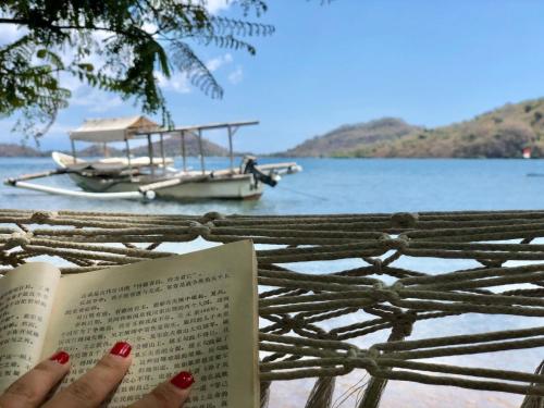 a woman reading a book with a boat in the water at Palmyra Indah Bungalows in Sekotong