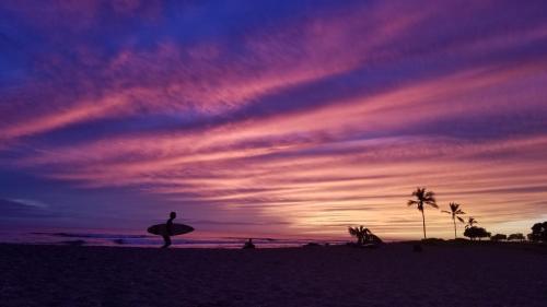 a person holding a surfboard on a beach with a sunset at DOWNTOWN Hawaiian Theme Condo with Hot Tub, Pool & Beach - Kona Islander Inn in Kailua-Kona