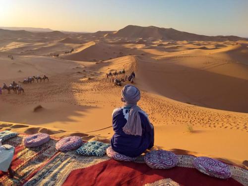 a woman sitting in the desert watching a camel caravan at OVERNIGHT BERBER CAMP in Merdani