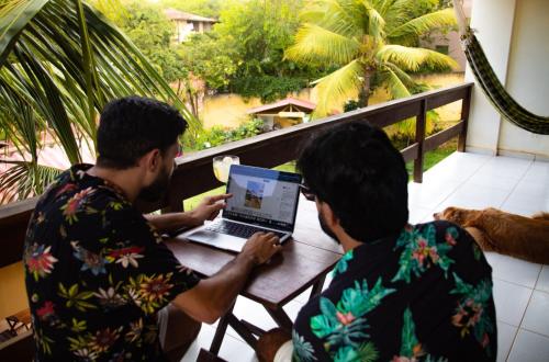 twee mannen aan een tafel met een laptop op een balkon bij Iduna Hostel in Pipa